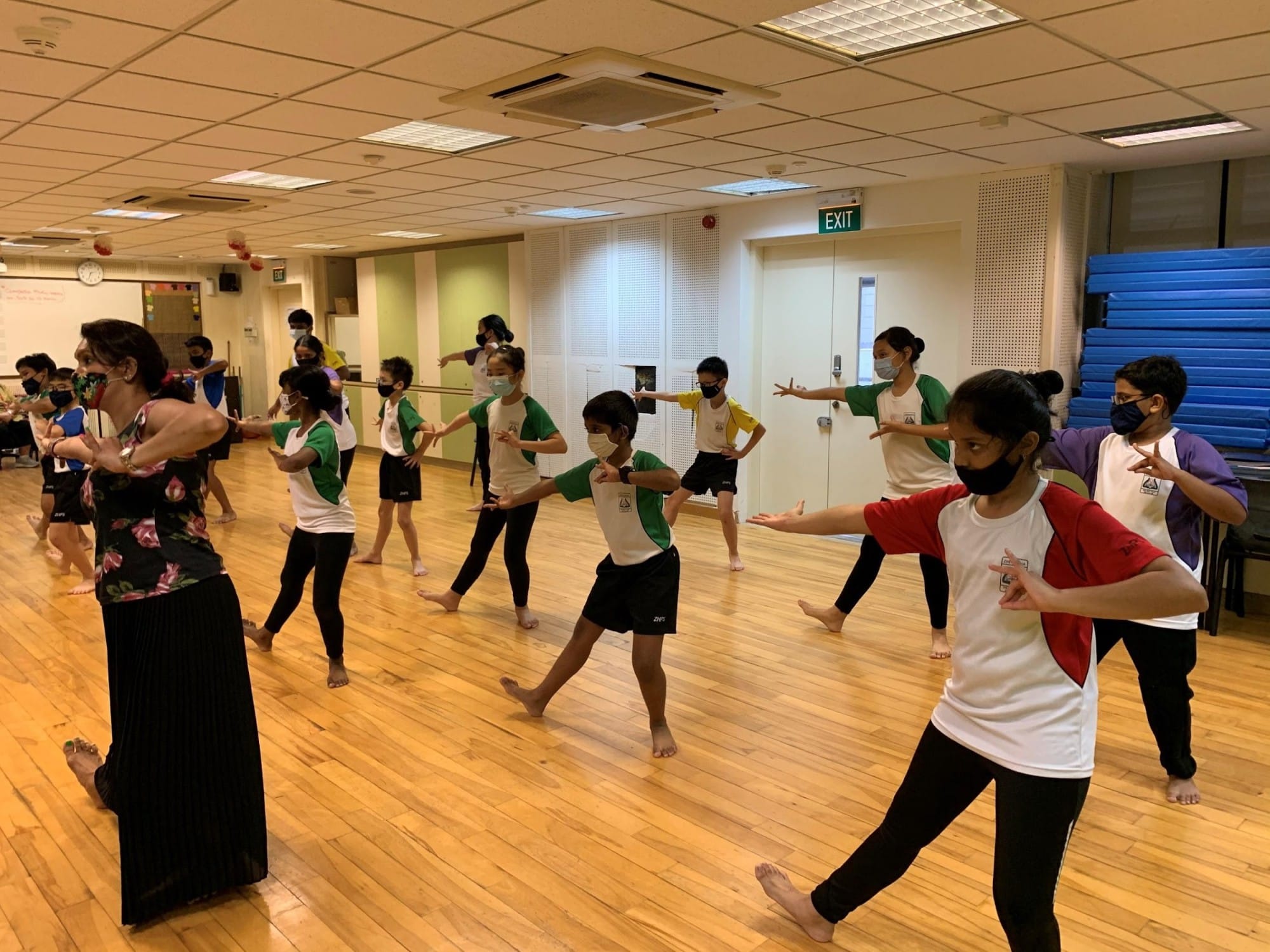 Children wearing masks practice a dance routine with an adult instructor in a wooden-floored indoor studio.